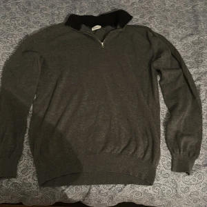 Mörkgrå halvzip-tröja från John Henric - Mörkgrå stickad halv-zip tröja från John Henric. Tröjan har långa ärmar och ribbade muddar vid ärmslut och krage. Den är i storlek m men sitter som s.