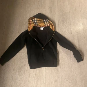 Svart hoodie med mönstrad huva från Burberry - Säljer en svart hoodie från Burberry med dragkedja och klassiskt rutigt foder i huvan. Jackan har långa ärmar, fickor framtill och guldfärgade detaljer på snörena. Perfekt för dig som gillar stilrena plagg med en twist.den är aldrig använd 
