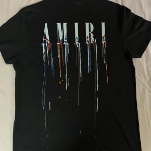 Svart AMIRI t-shirt med färgstänk - Svart t-shirt från AMIRI med stort tryck på ryggen och mindre tryck fram. Trycket består av AMIRI-loggan och färgstänk i blått, vitt, orange och brunt som rinner ner. Klassisk passform och rund hals. Perfekt för dig som gillar streetwear med edge.