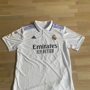 Real Madrid Rüdiger 22 Adidas tröja - Säljer en officiell Real Madrid matchtröja med Rüdiger 22 på ryggen. Tröjan är vit med lila Adidas-ränder på axlarna, klubbmärke och sponsortryck framtill. Klassisk krage och korta ärmar. Tillverkad i lätt och ventilerande polyester. Väldigt bra skick lite smutsig. Pris går att diskutera.