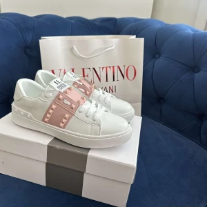 Valentino Rockstud sneakers vit/rosa - Snygga Valentino Rockstud sneakers i vitt skinn med breda ljusrosa band och ikoniska nitar. Klassisk rund tå, platt sula och vita snören. Perfekta för dig som vill sticka ut med en lyxig och trendig look.