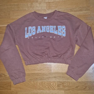 Brun croppad sweatshirt Los Angeles Lindex - Säljer en brun croppad sweatshirt från Lindex med trycket 'Los Angeles California' i vitt och ljusblått. Tröjan har långa ärmar, resår i midjan och är tillverkad i mjuk bomull. Perfekt för en chill och trendig look.