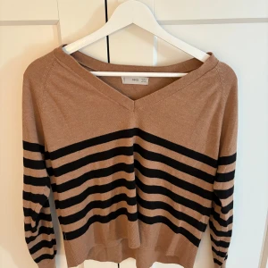 Randig beige tröja med v-ringning  - Snygg beige tröja från mango med svarta horisontella ränder och v-ringad hals. Tröjan har lång ärm och är i ett mjukt, stickat material. Perfekt för dig som gillar en avslappnad men ändå stilren look.