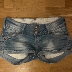 Blå lågmidjade jeansshorts - Snygga ljusblå jeansshorts från Mileno med slitna detaljer och fransig kant. Shortsen har 3 dekorativa knappar framtill, klassiska fickor och coola sömmar. Perfekta för sommaren och ger en avslappnad vibe till din outfit.