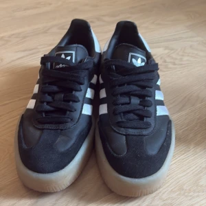 Adidas Samba svarta sneakers - Klassiska Adidas Samba sneakers i svart med vita ränder och vit häl. Skorna har snörning, platt sula i gummi och ovandel i skinn och mocka. Perfekta för dig som gillar retrostil och streetwear.