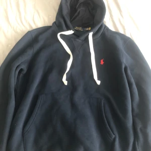 Mörkblå hoodie från Polo Ralph Lauren - Snygg mörkblå hoodie från Polo Ralph Lauren med vit snörning och klassisk röd logga broderad på bröstet. Tröjan har huva, känguruficka och långa ärmar. Perfekt för en chill och stilren look. Knappt använd och bra skick!