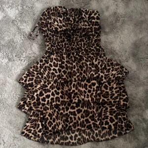 Leopardmönstrad volangklänning - Säljer en kort, axelbandslös klänning med leopardmönster och flera volanger. Klänningen har smockad överdel och tunna axelband som kan knytas. Perfekt för dig som vill sticka ut med ett djurmönstrat plagg! Den är onesize och passar på dig med storlekarna (xs, s, m, l) köpt i på en gatuaffär i Spanien! 