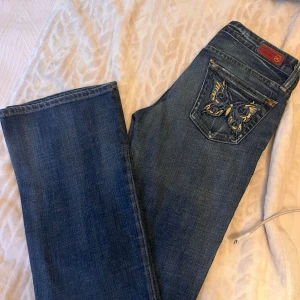 Blå jeans från AG Adriano Goldschmied - Snygga blå jeans från AG Adriano Goldschmied med broderad fjäril i gult och blått på bakfickan. Klassisk femficksmodell med utsvängda ben och coola slitningar framtill. Jeansen har normal passform och är tillverkade i mjukt bomullsjeans-tyg.