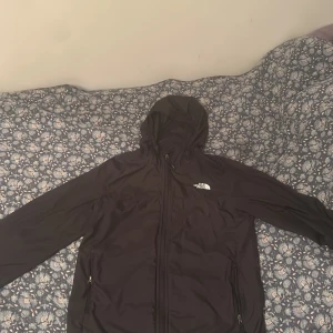 Svart vindjacka från The North Face - Svart vindjacka från The North Face med huva och dragkedja framtill. Jackan har två sidofickor med dragkedja och en stilren logga på bröstet. Perfekt för blåsiga dagar och enkel att matcha med allt.
