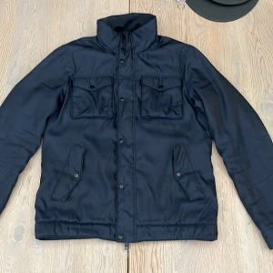 Marinblå Field jacket  från J.Lindeberg - Snygg marinblå field jacket från J.Lindeberg med hög krage, fyra fickor framtill och dragkedja med tryckknappar. Jackan har en stilren look och är perfekt för kyliga dagar. Materialet är syntet och jackan har en normal passform med långa ärmar.