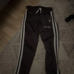 Palm Angels byxor - Svarta mjukisbyxor från Palm Angels med vita sidoränder och logga på låret. Byxorna har snörning i midjan och dragkedja vid fickorna. Perfekta för en chill och sportig look. 