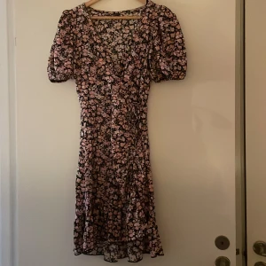 Blommig klänning från Gina Tricot - Supersöt blommig klänning från Gina Tricot i svart med rosa och vita blommor. Klänningen har v-ringning, puffiga korta ärmar och volang nertill. Figurnära passform och omlottkänsla framtill. Perfekt för vår och sommar! Använd 1 gång 