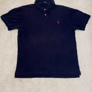Blå pikétröja Polo Ralph Lauren  - Klassisk blå pikétröja från Polo Ralph Lauren i slim fit-modell. Tröjan har krage, två knappar och den ikoniska röda broderade loggan på bröstet. Perfekt för en clean och stilren look.