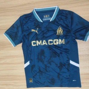 Marseille blå matchtröja Puma - Snygg blå fotbollströja från Olympique de Marseille med guldiga detaljer och logga. Kortärmad modell med ljusblå och vita inslag på krage och ärmslut. Tillverkad i lätt och ventilerande funktionsmaterial, perfekt för träning eller match.