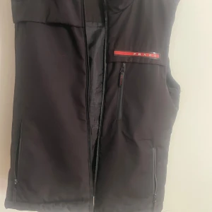 Svart dunväst från Prada - Svart dunväst från Prada med klassisk röd logga på bröstet. Västen har flera dragkedjeförsedda fickor och en stilren, minimalistisk design. Tillverkad i syntetmaterial och perfekt för lager-på-lager-stil. Passar dig som gillar exklusiva streetwear-vibbar. 