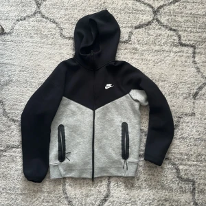 Nike tech svart och grå hoodiejacka M - Nike hoodiejacka i svart och ljusgrått med dragkedja hela vägen, två fickor med svarta detaljer och huva. Snygg sportig look med vit Nike-logga på bröstet. Perfekt för chill eller träning. Priser går alltid att jobba på. 