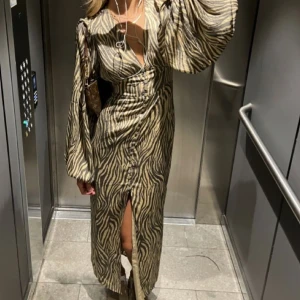 Zebramönstrad långklänning med slits - Snygg långklänning med zebramönster i beige och svart. Klänningen har v-ringning, långa vida ärmar och en slits framtill. Knappar längs framsidan ger en extra detalj. Perfekt för dig som vill sticka ut med en trendig look.