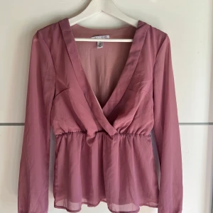Rosa omlottblus från NLY Trend - Säljer en superfin rosa blus från NLY Trend i omlottmodell med djup v-ringning och markerad midja. Blusen har långa ärmar och är gjord i ett tunt, lätt material som ger ett luftigt intryck. Perfekt att styla med jeans eller kjol för en trendig look.