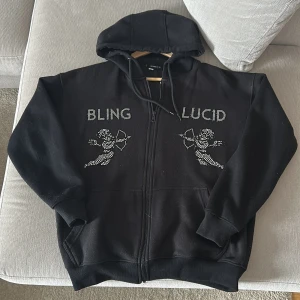 Svart hoodie med strasstenar från Blinglucid - Svart hoodie från Blinglucid i storlek XS med huva och dragkedja. Framsidan har coola strasstenar som bildar texten 'BLING LUCID' och två änglar med pilbåge. Mjuk bomullskänsla och klassisk passform, perfekt för dig som gillar statement-plagg. PRIS KAN SJÄLVKLART DISKUTERAS 