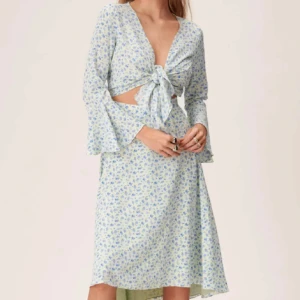 Blommig ljusblå omlottklänning med knyt - Superfin ljusblå klänning med små blommor, v-ringning och knyt framtill. Klänningen har långa ärmar med lätt utsvängda ärmslut och cut-out detaljer vid midjan. Perfekt för sommarens alla tillfällen!