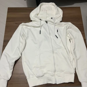 Vit hoodie från Polo Ralph Lauren - Snygg vit hoodie från Polo Ralph Lauren med dragkedja och klassisk logga på bröstet. Tröjan har huva med snörning, känguruficka och ribbade muddar. Perfekt för en chill och stilren look. 