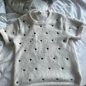 Vit stickad tröja med svarta hjärtan - Supersöt vit stickad tröja med korta ärmar och små svarta hjärtan över hela. Mjuk och fluffig känsla, perfekt för dig som gillar gulliga detaljer. Har ett litet hål som går att fixas💕💕