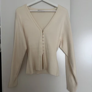 Beige kofta med knappar från Gina Tricot - Superfin beige kofta från Gina Tricot i storlek XS. Den har v-ringning, långa ärmar och snygga tygklädda knappar framtill. Koftan är i mjukt stickat material och har en liten slits nedtill framtill. Perfekt för lager-på-lager och stilrena outfits. Knappt använd. Finns en sminkfläck som går bort i tvätten.