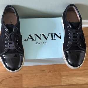 Svarta sneakers från Lanvin med lackad tå - Snygga svarta sneakers från Lanvin med ovandel i mocka och en glansig lackad tå. Skorna har vita sulor, svarta snören och klassisk låg profil. Perfekta för dig som gillar stilrena och exklusiva detaljer.