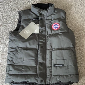 Grå Canada gosse väst pris kan diskuteras  - Säljer en helt ny, oanvänd Canada Goose väst i grått. Klassisk modell med logotyp på bröstet, stora fickor och hög krage. Västen stängs med knappar och dragkedja och passar perfekt för lager-på-lager under vår höst och Sveriges sommar dagar. Storlek M, men funkar även för dig som har S eller L beroende på hur du vill att den ska sitta. Allting som är med i bilden ingår med västen, påse och tags. Nypris betydligt högre — pris kan diskuteras vid snabb affär.