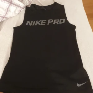 Svart ärmlös Nike Pro träningstopp - Svart ärmlös topp från Nike Pro med grå logga framtill och liten swoosh nertill. Tillverkad i ett lätt och stretchigt material som andas, perfekt för intensiva träningspass. Snygg omlott-detalj baktill ger extra stilpoäng och rörelsefrihet. Baksidan har ett luftigare material