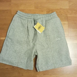 Grå sweatshorts från Cortiez  - Grå sweatshorts med broderad Cortiez-logga framtill och HMP-märke med röda stjärnor på bakfickan i storlek S. Mjuk resår i midjan med snörning och praktiska sidofickor. Perfekta för chill dagar eller när du vill ha en avslappnad look. 