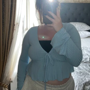 Ljusblå ribbad omlottopp med knyt - Supersöt ljusblå omlottopp med ribbad struktur och långa ärmar. Toppen har v-ringning, knyt framtill och lätt croppad passform med vågig kant nertill. Perfekt för dig som gillar en feminin och trendig stil.