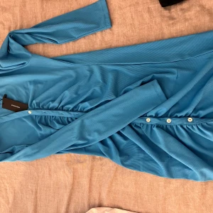 Blå ribbad klänning från Vero Moda - Supersnygg blå ribbad klänning från Vero Moda med v-ringning, långa ärmar och knappar framtill. Klänningen har en figurnära passform och fina rynkade detaljer vid midjan. Perfekt för dig som vill sticka ut med färg och stil.