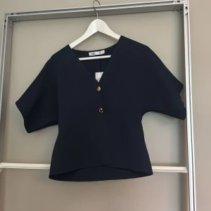 Mörkblå blus med v-ringning från Zara - Snygg mörkblå blus från Zara med v-ringning och två guldiga knappar framtill. Modellen är boxig med korta, vida ärmar och lätt strukturerat tyg. Perfekt för dig som gillar stilrena och moderna plagg med en twist. Aldrig använd!