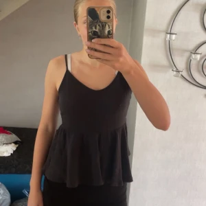 Svart/mörk brunt ribbad linnetopp med volang - Svart/mörkt brunt ribbad linnetopp med smala axelband och volang nertill. Toppen har v-ringad hals och är figurnära upptill med en lösare passform nedtill. Några fläckar som men allra högsta sannolikhet går bort i tvätten. Även ett litet hål vid höger band, bak. Inget som syns tydligt🤎