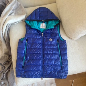 Blå dunväst med huva från Moncler - Snygg blå dunväst från Moncler med quiltad design. Västen har dragkedja framtill, två fickor med dragkedja och en liten bröstficka med Moncler-logga. Insidan är turkos och materialet är glansigt och lätt. 