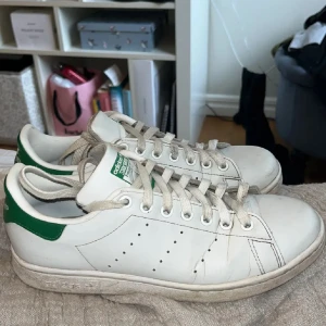 Adidas Stan Smith vita sneakers - Klassiska Adidas Stan Smith sneakers i vitt skinn med gröna detaljer på häl och plös. Skorna har rund tå, snörning och perforerade ränder på sidorna. Perfekta för en clean och sportig look.