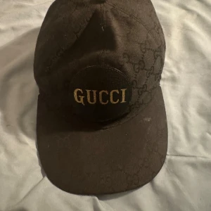 Gucci keps svart  - Unik Snygg svart keps från Gucci med klassiskt GG-mönster över hela kepsen och en rund patch framtill med guldig GUCCI-text. Kepsen är tillverkad i bomull och har en stilren, böjd skärm. Perfekt för dig som vill ha en exklusiv streetstyle-look, 
