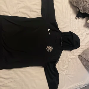 Svart AIK Nike hoodie XL Dri-Fit - Svart hoodie från Nike med AIK-emblem på bröstet och klassisk vit swoosh. Tillverkad i Dri-Fit-material som andas, perfekt för fotboll eller chill. Långärmad modell med huva och sportig vibe. Passar dig som vill visa AIK-stolthet på träning eller i vardagen.