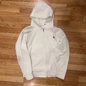 Vit hoodie från Ralph Lauren - Vit hoodie från Ralph Lauren med dragkedja framtill och klassisk broderad logga på bröstet. Tröjan har huva, känguruficka och långa ärmar. Perfekt för en chill och stilren look.