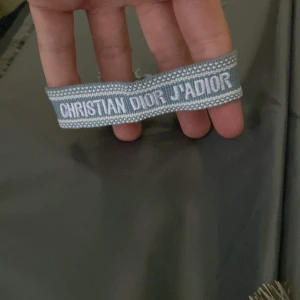 Blått Christian Dior J'Adior armband - Snyggt vävt armband från Christian Dior i ljusblått med vit brodyr där det står 'CHRISTIAN DIOR J'ADIOR'. Armbandet har justerbar knytning med tofsar i ändarna för en cool och avslappnad look.