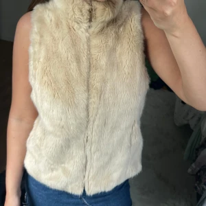 Beige pälsväst med dragkedja - Snygg och fluffig beige pälsväst med hög krage och dragkedja framtill. Västen har en rak passform och är perfekt att slänga över en hoodie eller långärmad tröja för en cool lager-på-lager-look.