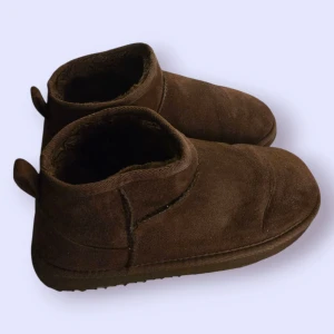 Bruna mockaboots från Carin Wester - Mysiga bruna boots i mocka från Carin Wester med mjukt foder på insidan. Skorna har rund tå, platt sula och låg profil, perfekta för kalla dagar. Klassisk design med dragflik bak och diskreta sömmar för en enkel men stilren look.