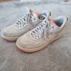 Nike Air Force 1 vita med röd sula - Nike Air Force 1 sneakers i vitt med detaljer i läder och canvas. Skorna har en klassisk siluett, snörning och röd yttersula som ger en snygg kontrast. Tungan har röd Nike Air-logga och små röda detaljer på sidan. Perfekta för dig som gillar stilrena sneakers med en twist.