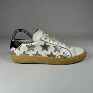 Saint Laurent sneakers med silverstjärnor - Unika vita sneakers från Saint Laurent med metallic-silver stjärnor på sidorna och svart detalj vid hälen. Skorna har beige sula, snörning och är tillverkade i skinn. Perfekta för dig som vill sticka ut med en cool och trendig look.