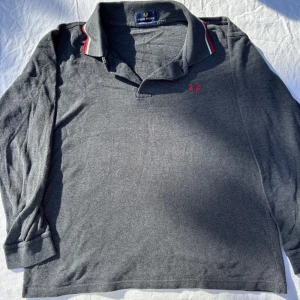 Grå långärmad pikétröja Fred Perry - Snygg grå långärmad pikétröja från Fred Perry i 100% bomull. Klassisk krage med röda och vita ränder, knappar framtill och ikonisk röd logga broderad på bröstet. Perfekt för en stilren och avslappnad look.
