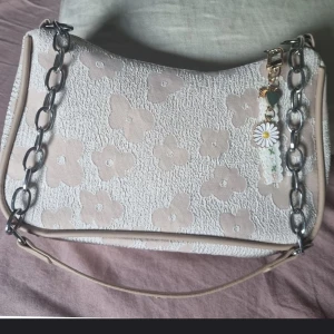 Axelväska  - En söt blommigt axelväska i kaki färg och en silver kedja som axelrem. Väskan har en liten berlock med en blomma och andra detaljer. Insidan är fodrad i svart.  Mått 14 X 22 X 7 cm