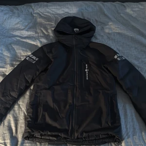 Svart Sail Racing vindjacka med huva - Svart jacka från Sail Racing med huva och dragkedja. Jackan är vattentät, vindtät och andas, med logga på bröstet och ärmarna. Snygg och sportig design. Flera praktiska fickor och justerbar nederkant.