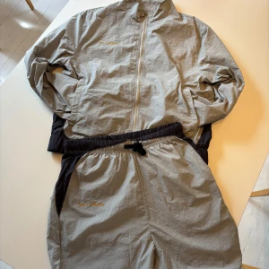 Grå tracksuit från Sens Collective - Grå tracksuit från Sens Collective med gul broderad logga på båda plaggen. Jackan har hel dragkedja framtill, ståkrage och svarta detaljer längs midjan. Lätt och smidig modell i syntetmaterial, perfekt för lager-på-lager. Snygg och enkel att matcha till olika outfits.