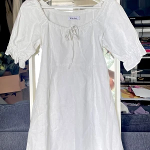 Vit mini dress från With Jean - Supersöt vit kortklänning från With Jean i linne med puffärmar, knytning fram och rosett i ryggen. Klänningen har en luftig passform och är perfekt för varma dagar. Enkel och stilren design som känns både trendig och tidlös. Material är linne och bomull och klänningen är helt oanvänd pga fel passform. 